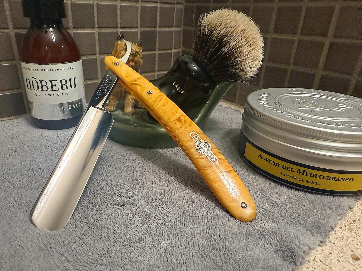 On a gray microfiber towel a straight razor with blade open about 45 degrees leans next to a bottle of aftershave and a jar of shave soap. A shave brush rests bristles-up in a green Wade ceramic pipe rest that features a brown Cairn Terrier.