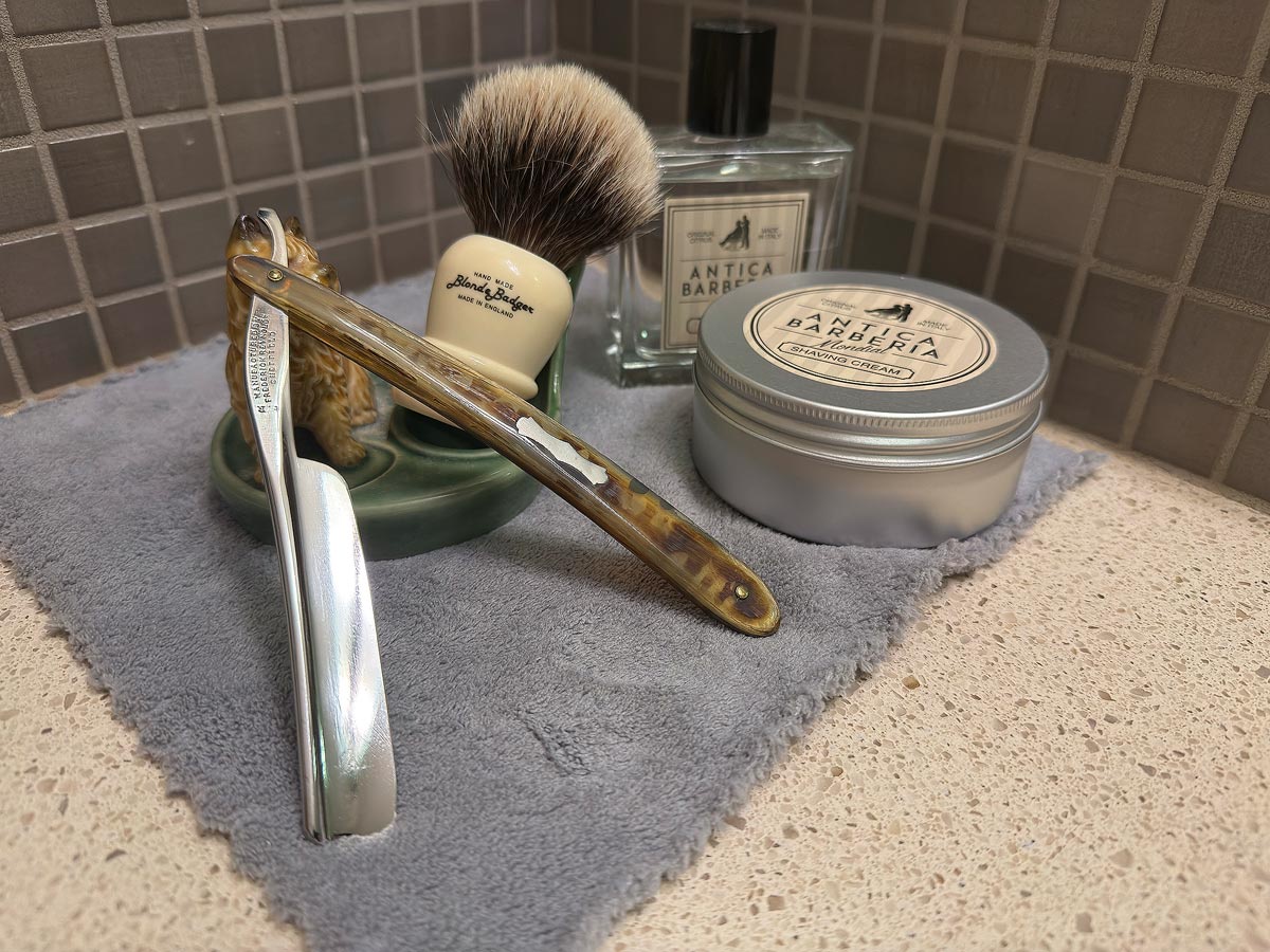 On a gray microfiber towel a straight razor with blade open about 45 degrees leans next to a bottle of aftershave and a jar of shave soap. A shave brush rests bristles-up in a green Wade ceramic pipe rest that features a brown Cairn Terrier.