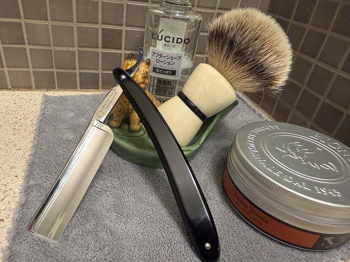 On a gray microfiber towel a straight razor with blade open about 45 degrees leans next to a bottle of aftershave and a jar of shave soap. A shave brush rests bristles-up in a green Wade ceramic pipe rest that features a brown Cairn Terrier.