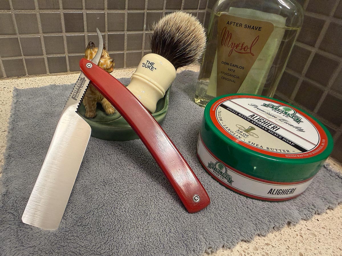 On a gray microfiber towel a straight razor with blade open about 45 degrees leans next to a bottle of aftershave and a jar of shave soap. A shave brush rests bristles-up in a green Wade ceramic pipe rest that features a brown Cairn Terrier.