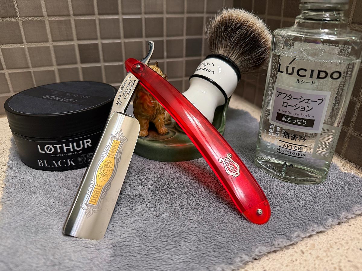 On a gray microfiber towel a straight razor with blade open about 45 degrees leans next to a bottle of aftershave and a jar of shave soap. A shave brush rests bristles-up in a green Wade ceramic pipe rest that features a brown Cairn Terrier.