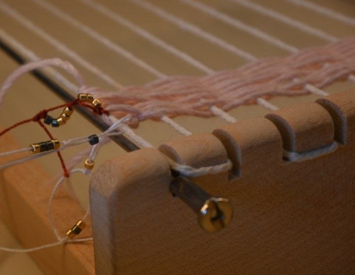 foto de un telar en el que hay anudados hilos blanco, rojo y rosa con cuentas doradas y azules
