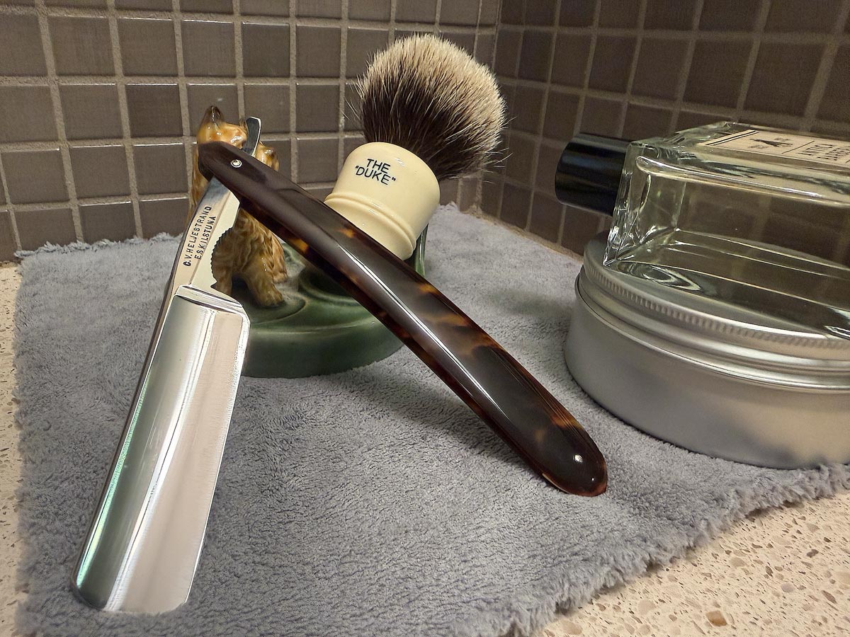 On a gray microfiber towel a straight razor with blade open about 45 degrees leans next to a bottle of aftershave and a jar of shave soap. A shave brush rests bristles-up in a green Wade ceramic pipe rest that features a brown Cairn Terrier.