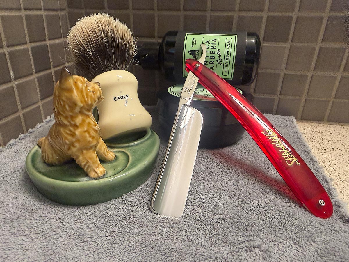 On a gray microfiber towel a straight razor with blade open about 45 degrees leans next to a bottle of aftershave and a jar of shave soap. A shave brush rests bristles-up in a green Wade ceramic pipe rest that features a brown Cairn Terrier.