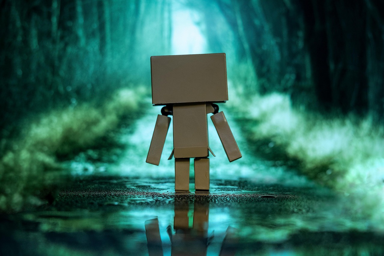 A cardboard box robot faces away from the camera and walks down a wet, wooded road.