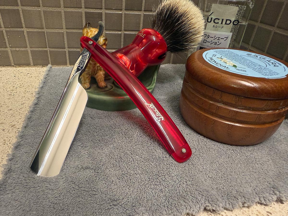 On a gray microfiber towel a straight razor with blade open about 45 degrees leans next to a bottle of aftershave and a jar of shave soap. A shave brush rests bristles-up in a green Wade ceramic pipe rest that features a brown Cairn Terrier.