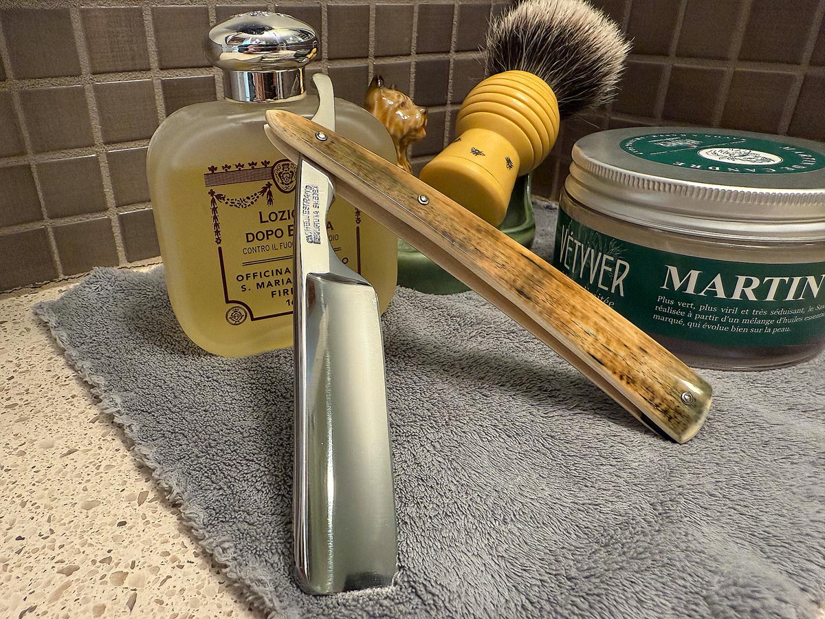 On a gray microfiber towel a straight razor with blade open about 45 degrees leans next to a bottle of aftershave and a jar of shave soap. A shave brush rests bristles-up in a green Wade ceramic pipe rest that features a brown Cairn Terrier.