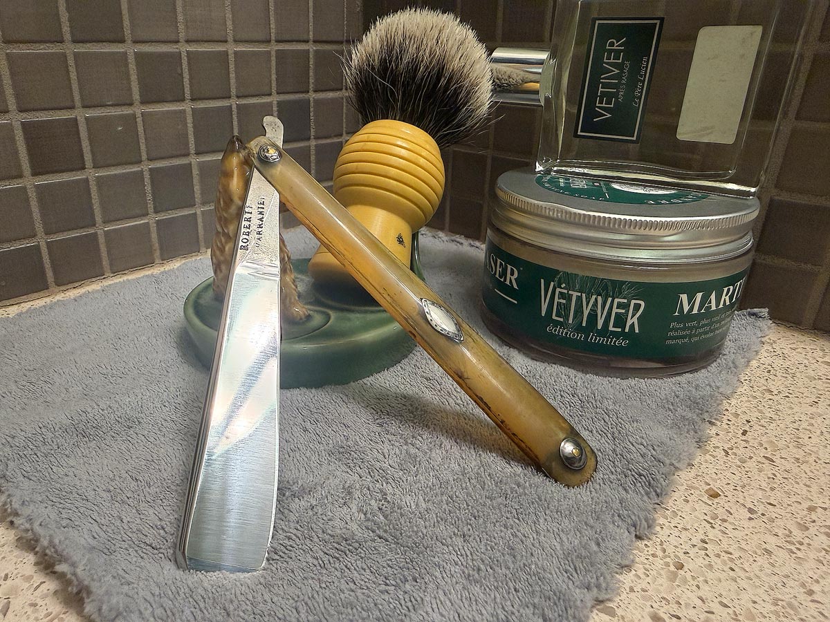 On a gray microfiber towel a straight razor with blade open about 45 degrees leans next to a bottle of aftershave and a jar of shave soap. A shave brush rests bristles-up in a green Wade ceramic pipe rest that features a brown Cairn Terrier.