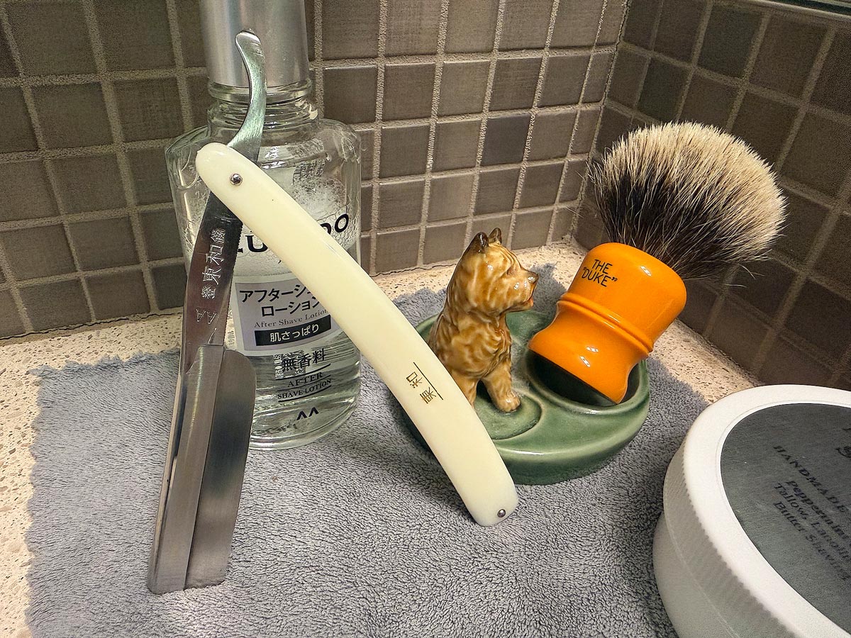 On a gray microfiber towel a straight razor with blade open about 45 degrees leans next to a bottle of aftershave and a jar of shave soap. A shave brush rests bristles-up in a green Wade ceramic pipe rest that features a brown Cairn Terrier.