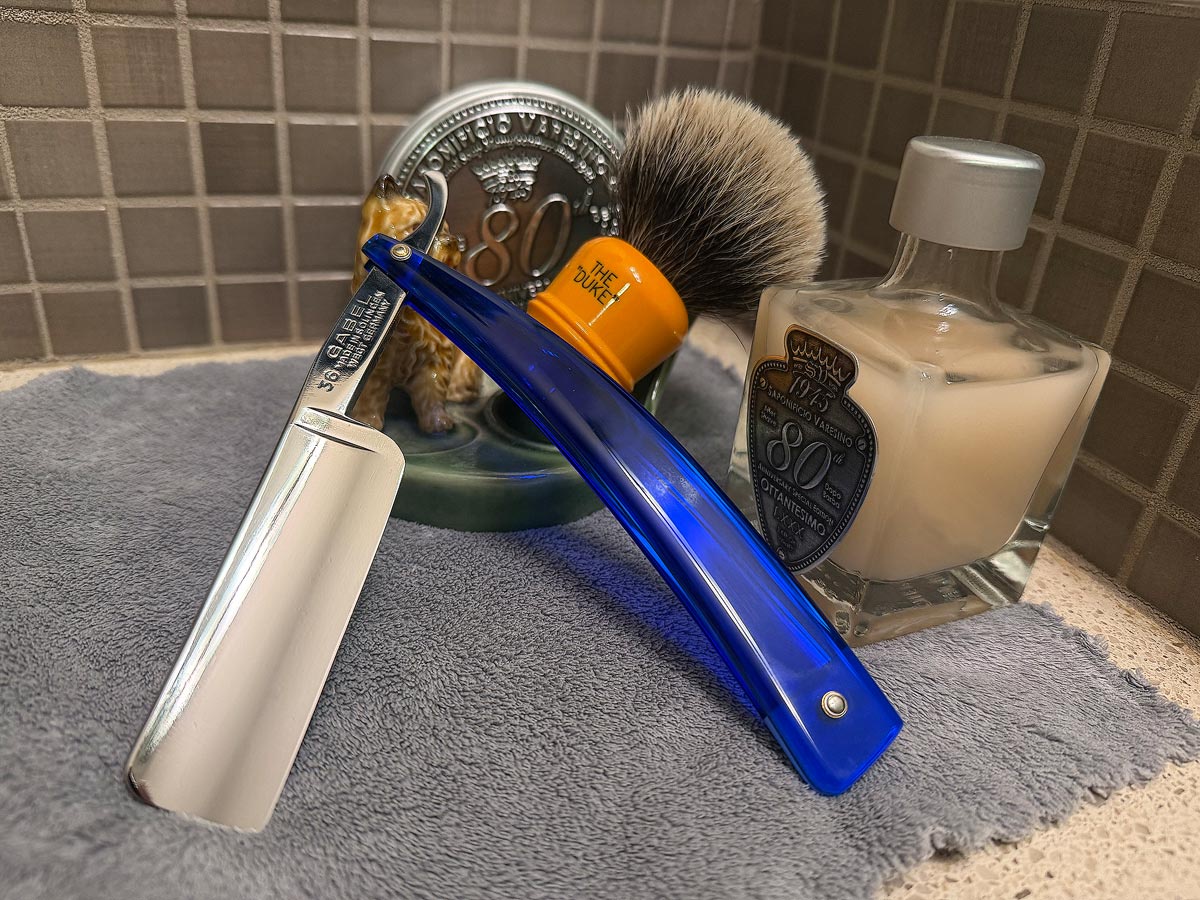 On a gray microfiber towel a straight razor with blade open about 45 degrees leans next to a bottle of aftershave and a jar of shave soap. A shave brush rests bristles-up in a green Wade ceramic pipe rest that features a brown Cairn Terrier.