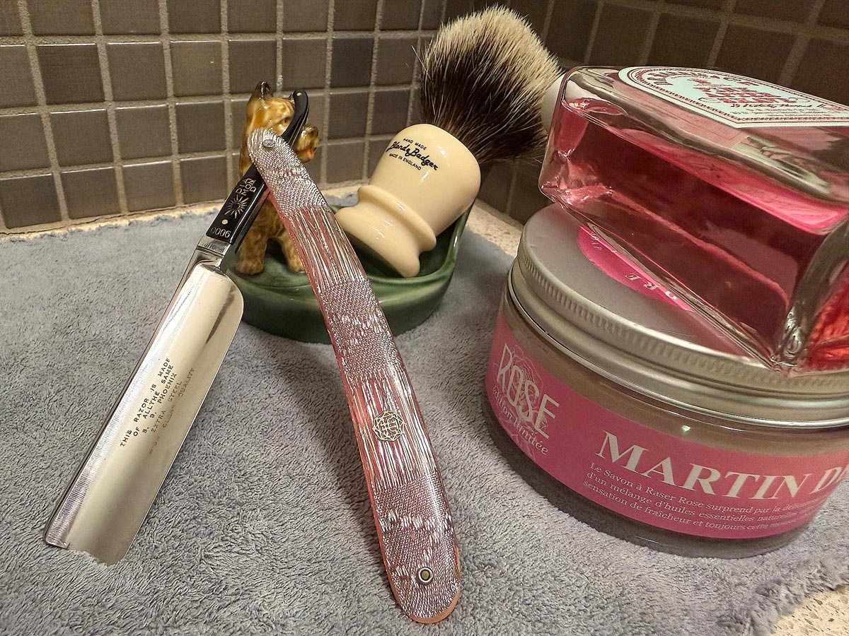 On a gray microfiber towel a straight razor with blade open about 45 degrees leans next to a bottle of aftershave and a jar of shave soap. A shave brush rests bristles-up in a green Wade ceramic pipe rest that features a brown Cairn Terrier.