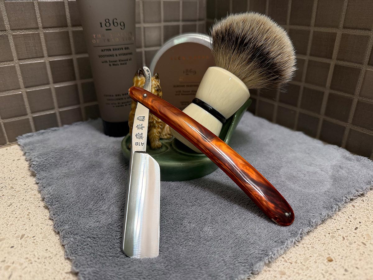 On a gray microfiber towel a straight razor with blade open about 45 degrees leans next to a bottle of aftershave and a jar of shave soap. A shave brush rests bristles-up in a green Wade ceramic pipe rest that features a brown Cairn Terrier.