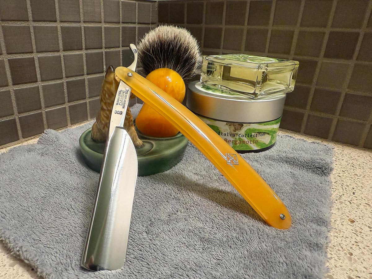 On a gray microfiber towel a straight razor with blade open about 45 degrees leans next to a bottle of aftershave and a jar of shave soap. A shave brush rests bristles-up in a green Wade ceramic pipe rest that features a brown Cairn Terrier.