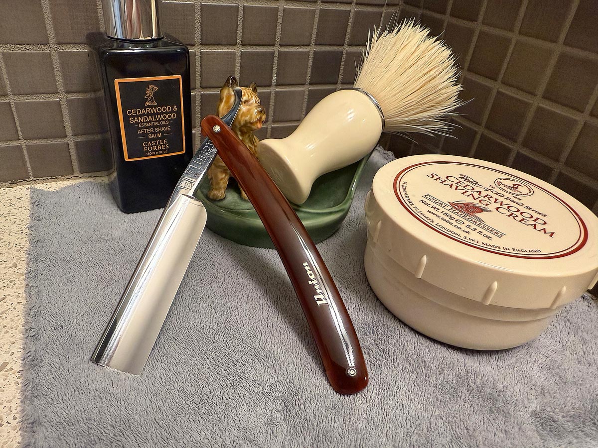 On a gray microfiber towel a straight razor with blade open about 45 degrees leans next to a bottle of aftershave and a jar of shave soap. A shave brush rests bristles-up in a green Wade ceramic pipe rest that features a brown Cairn Terrier.