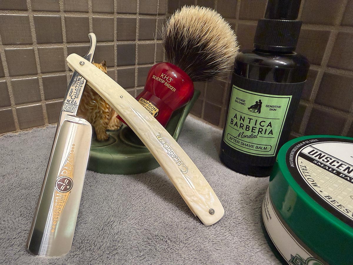 On a gray microfiber towel a straight razor with blade open about 45 degrees leans next to a bottle of aftershave and a jar of shave soap. A shave brush rests bristles-up in a green Wade ceramic pipe rest that features a brown Cairn Terrier.