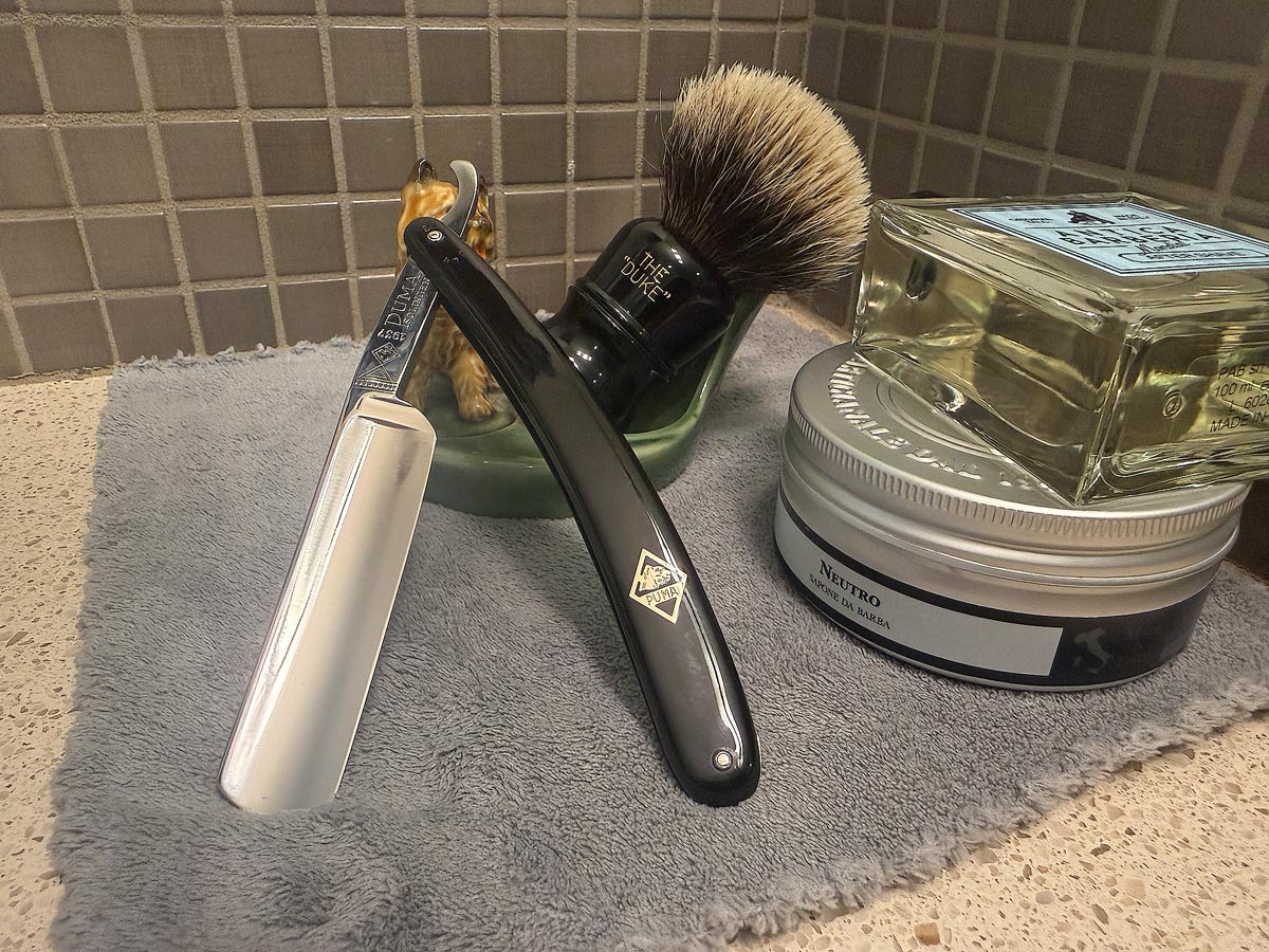 On a gray microfiber towel a straight razor with blade open about 45 degrees leans next to a bottle of aftershave and a jar of shave soap. A shave brush rests bristles-up in a green Wade ceramic pipe rest that features a brown Cairn Terrier.