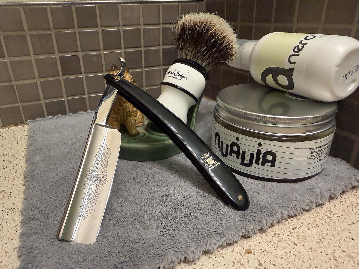 On a gray microfiber towel a straight razor with blade open about 45 degrees leans next to a bottle of aftershave and a jar of shave soap. A shave brush rests bristles-up in a green Wade ceramic pipe rest that features a brown Cairn Terrier.
