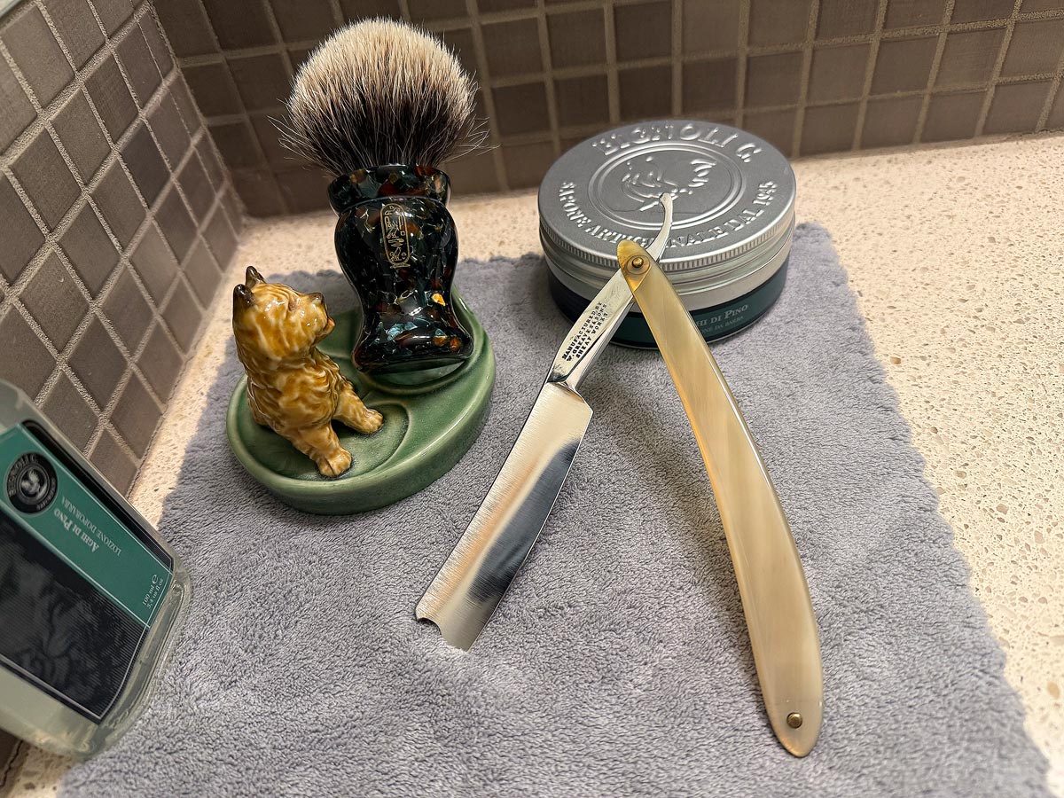 On a gray microfiber towel a straight razor with blade open about 45 degrees leans next to a bottle of aftershave and a jar of shave soap. A shave brush rests bristles-up in a green Wade ceramic pipe rest that features a brown Cairn Terrier.