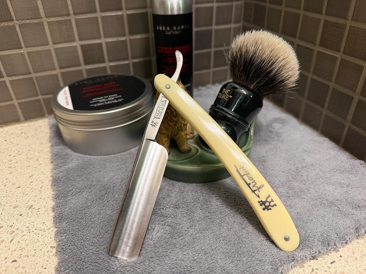 On a gray microfiber towel a straight razor with blade open about 45 degrees leans next to a bottle of aftershave and a jar of shave soap. A shave brush rests bristles-up in a green Wade ceramic pipe rest that features a brown Cairn Terrier.