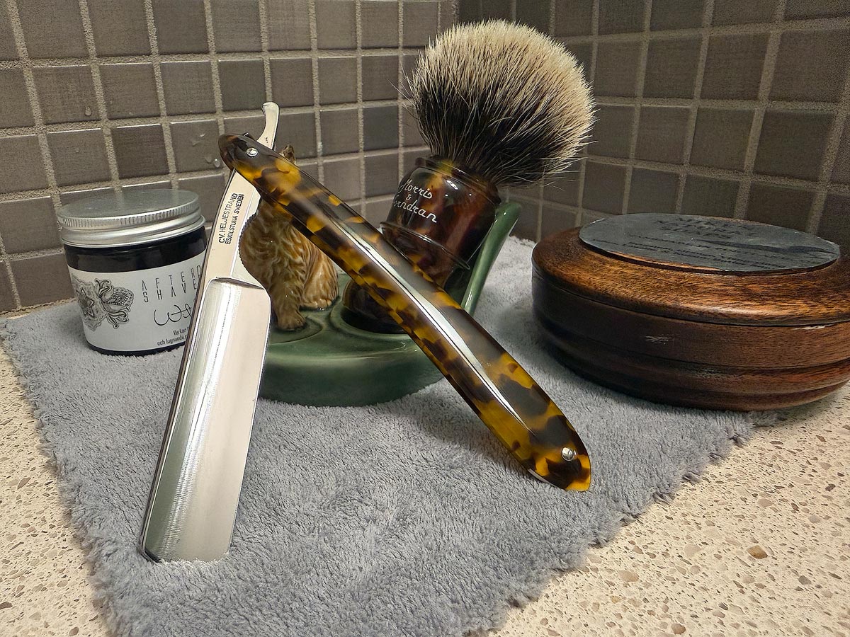 On a gray microfiber towel a straight razor with blade open about 45 degrees leans next to a bottle of aftershave and a jar of shave soap. A shave brush rests bristles-up in a green Wade ceramic pipe rest that features a brown Cairn Terrier.