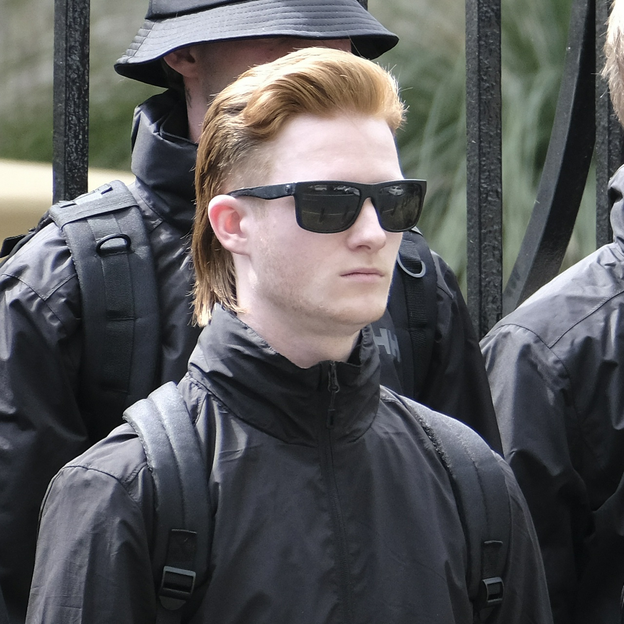A very pale, thin ginger with slicked-back shoulder length hair, small features and a large forehead wearing thick black sunglasses