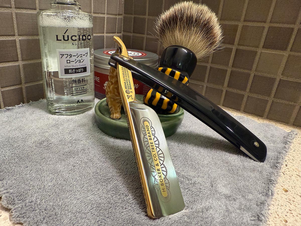 On a gray microfiber towel a straight razor with blade open about 45 degrees leans next to a bottle of aftershave and a jar of shave soap. A shave brush rests bristles-up in a green Wade ceramic pipe rest that features a brown Cairn Terrier.