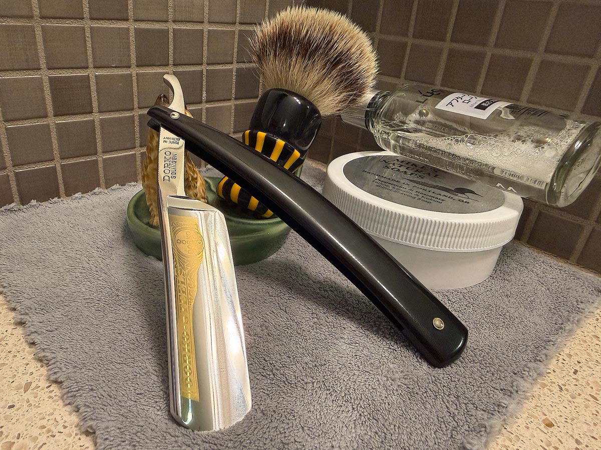 On a gray microfiber towel a straight razor with blade open about 45 degrees leans next to a bottle of aftershave and a jar of shave soap. A shave brush rests bristles-up in a green Wade ceramic pipe rest that features a brown Cairn Terrier.