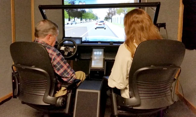 Two people sitting in office chairs in front of a full-size automobile cabin emulator. It houses a steering wheel, console, multiple dashboard screens, and a giant simulated windshield with a road and cars on a TV screen behind it.