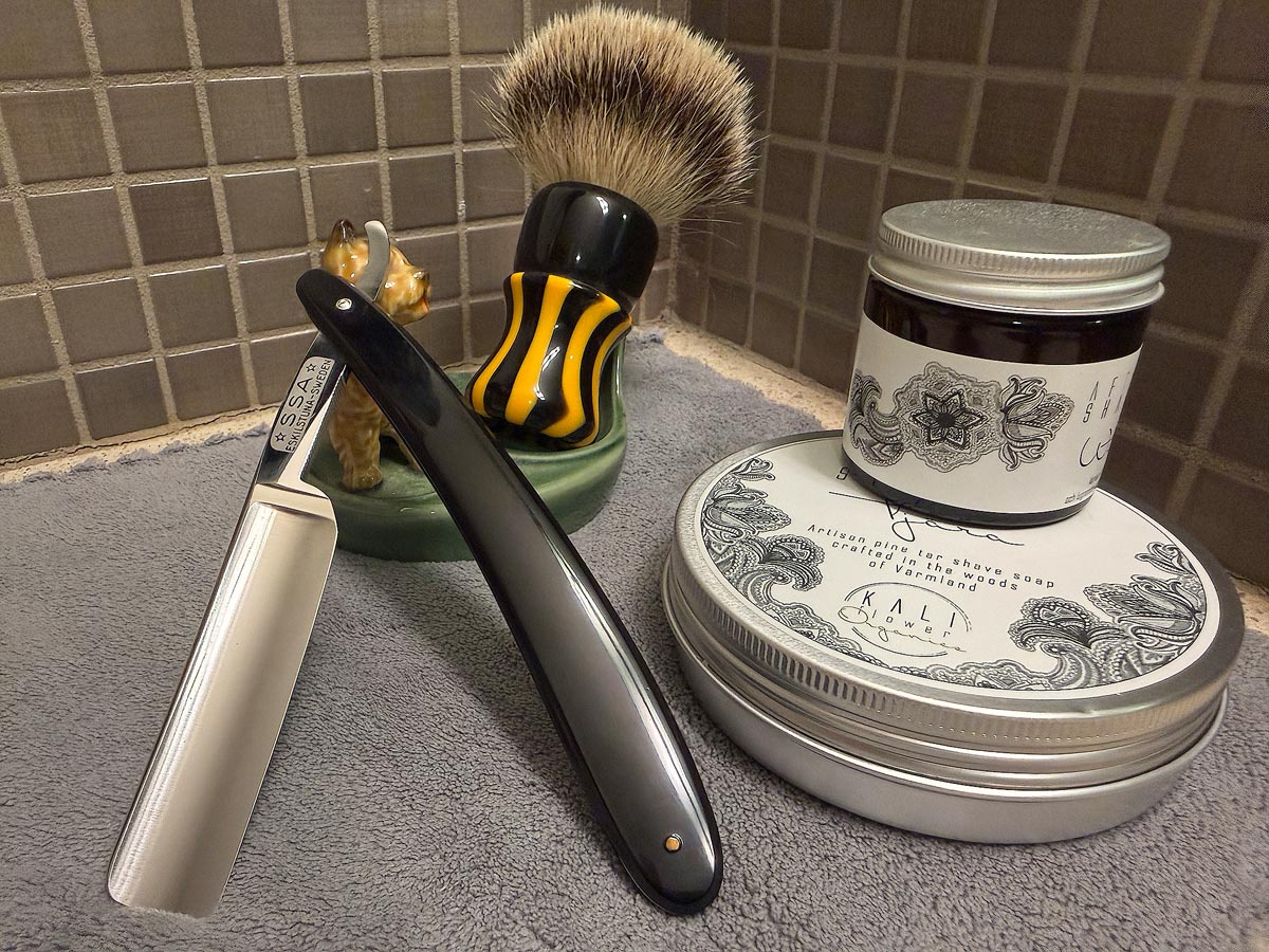 On a gray microfiber towel a straight razor with blade open about 45 degrees leans next to a bottle of aftershave and a jar of shave soap. A shave brush rests bristles-up in a green Wade ceramic pipe rest that features a brown Cairn Terrier.