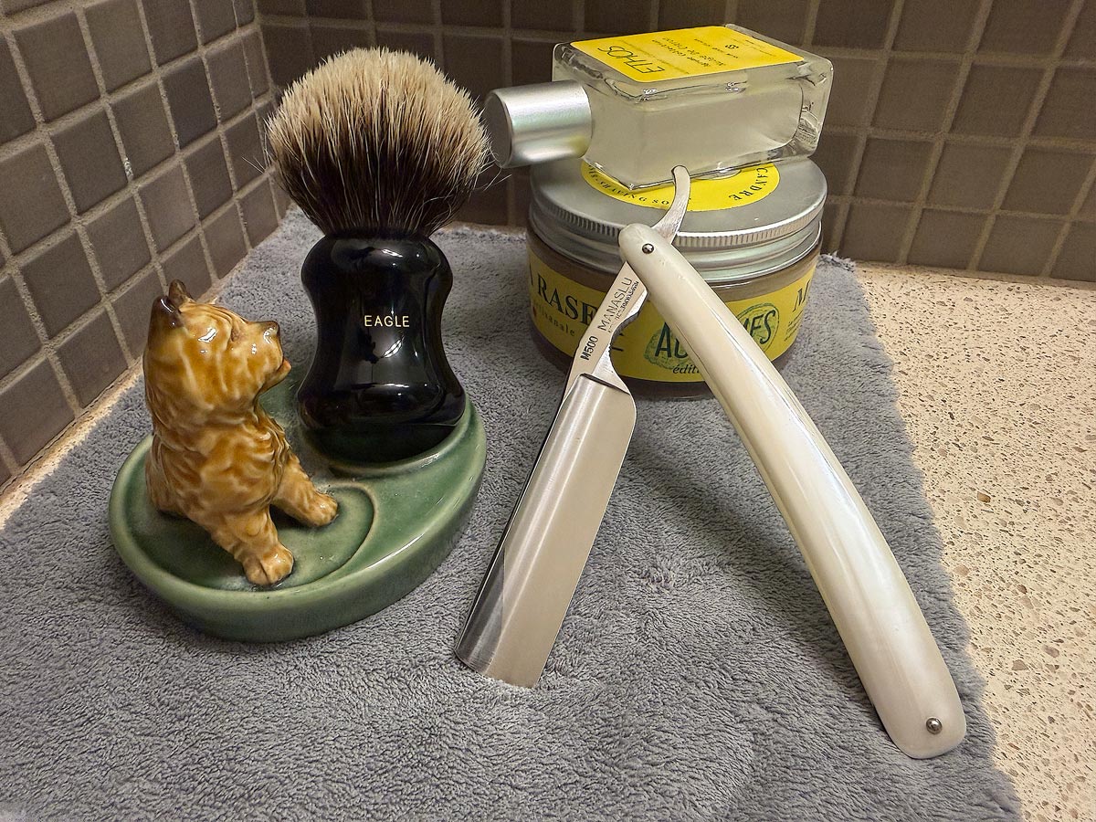 On a gray microfiber towel a straight razor with blade open about 45 degrees leans next to a bottle of aftershave and a jar of shave soap. A shave brush rests bristles-up in a green Wade ceramic pipe rest that features a brown Cairn Terrier.