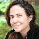 Naomi, gently smiling, long brown hair parted to the side in a pony tail, wearing a casual shirt, outside with some foliage behind her.