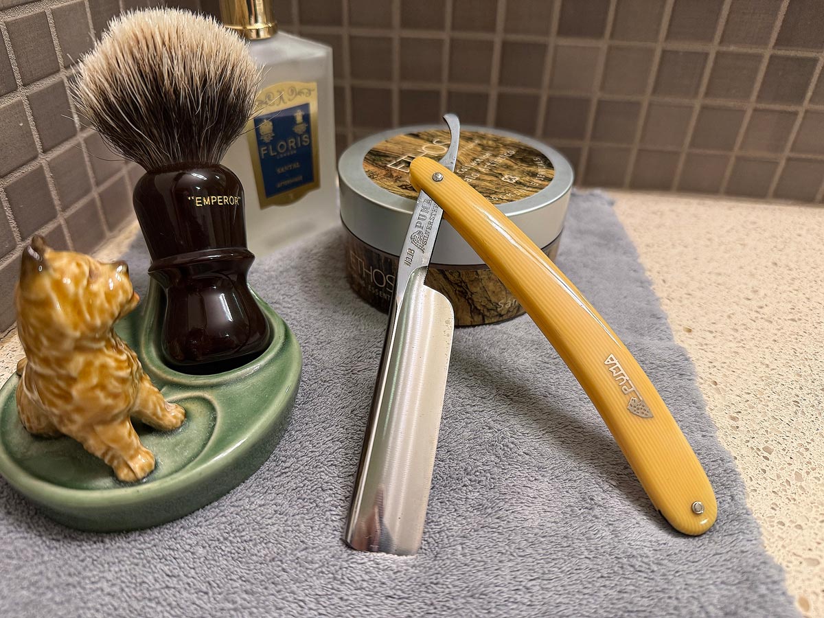On a gray microfiber towel a straight razor with blade open about 45 degrees leans next to a bottle of aftershave and a jar of shave soap. A shave brush rests bristles-up in a green Wade ceramic pipe rest that features a brown Cairn Terrier.