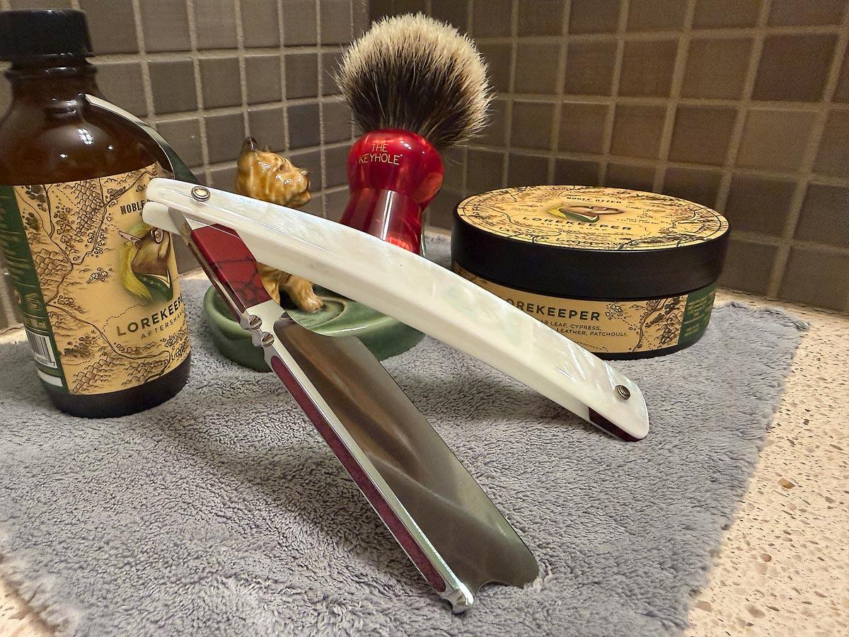 On a gray microfiber towel a straight razor with blade open about 45 degrees leans next to a bottle of aftershave and a jar of shave soap. A shave brush rests bristles-up in a green Wade ceramic pipe rest that features a brown Cairn Terrier.