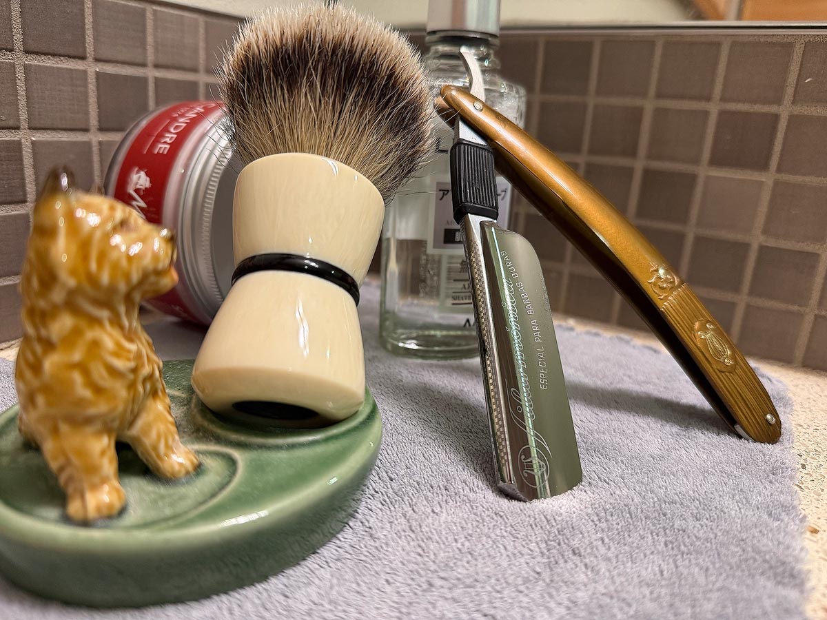 On a gray microfiber towel a straight razor with blade open about 45 degrees leans next to a bottle of aftershave and a jar of shave soap. A shave brush rests bristles-up in a green Wade ceramic pipe rest that features a brown Cairn Terrier.