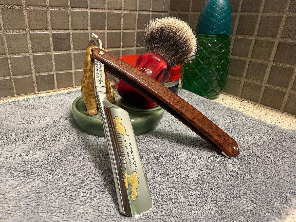 On a gray microfiber towel a straight razor with blade open about 45 degrees leans next to a bottle of aftershave and a jar of shave soap. A shave brush rests bristles-up in a green Wade ceramic pipe rest that features a brown Cairn Terrier.