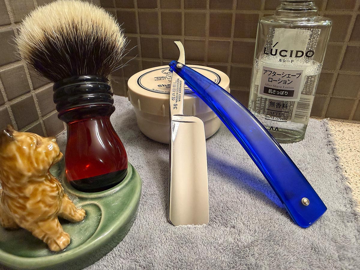 On a gray microfiber towel a straight razor with blade open about 45 degrees leans next to a bottle of aftershave and a jar of shave soap. A shave brush rests bristles-up in a green Wade ceramic pipe rest that features a brown Cairn Terrier.