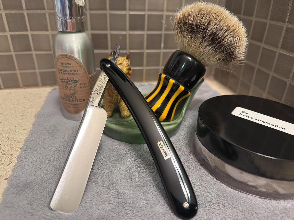 On a gray microfiber towel a straight razor with blade open about 45 degrees leans next to a bottle of aftershave and a jar of shave soap. A shave brush rests bristles-up in a green Wade ceramic pipe rest that features a brown Cairn Terrier.