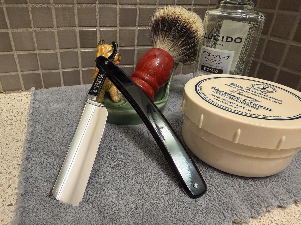 On a gray microfiber towel a straight razor with blade open about 45 degrees leans next to a bottle of aftershave and a jar of shave soap. A shave brush rests bristles-up in a green Wade ceramic pipe rest that features a brown Cairn Terrier.