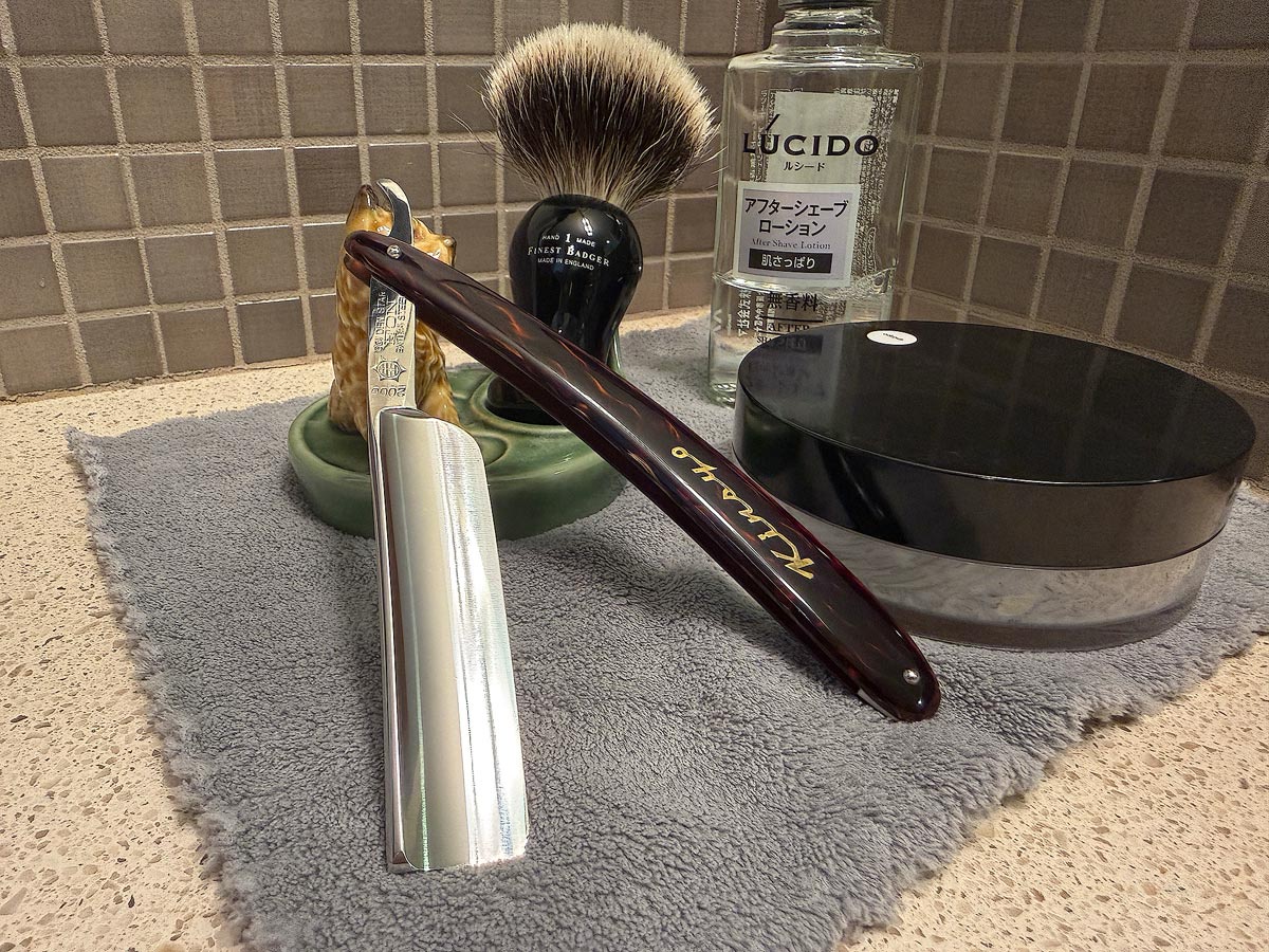 On a gray microfiber towel a straight razor with blade open about 45 degrees leans next to a bottle of aftershave and a jar of shave soap. A shave brush rests bristles-up in a green Wade ceramic pipe rest that features a brown Cairn Terrier.