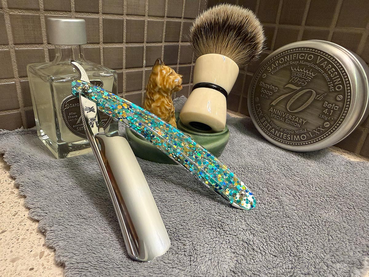 On a gray microfiber towel a straight razor with blade open about 45 degrees leans next to a bottle of aftershave and a jar of shave soap. A shave brush rests bristles-up in a green Wade ceramic pipe rest that features a brown Cairn Terrier.