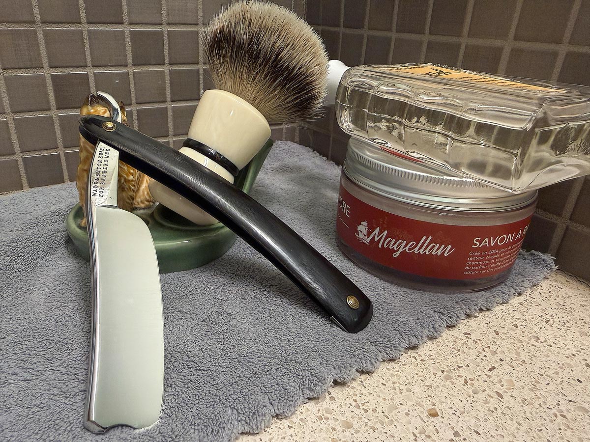 On a gray microfiber towel a straight razor with blade open about 45 degrees leans next to a bottle of aftershave and a jar of shave soap. A shave brush rests bristles-up in a green Wade ceramic pipe rest that features a brown Cairn Terrier.
