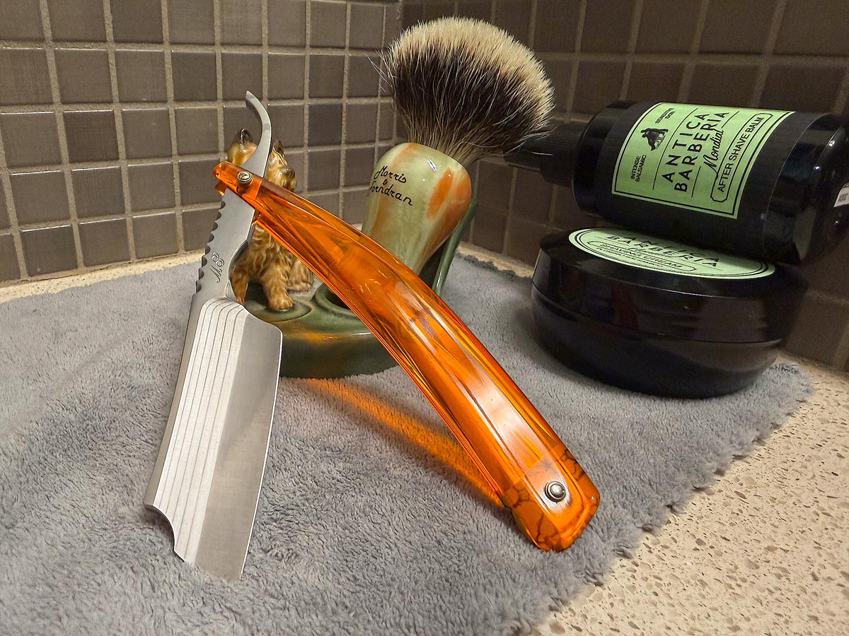 On a gray microfiber towel a straight razor with blade open about 45 degrees leans next to a bottle of aftershave and a jar of shave soap. A shave brush rests bristles-up in a green Wade ceramic pipe rest that features a brown Cairn Terrier.