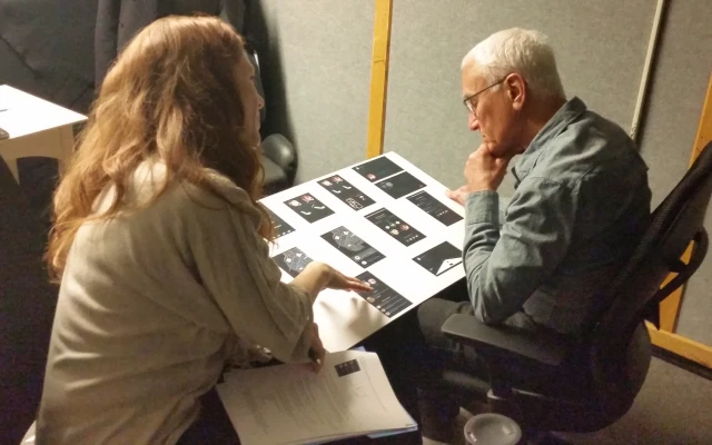 An older man in a dress shirt, looking over a giant board of user interface screens with his fist on his chin, deep in analytical thought. A woman with long red hair sits beside him with her hand over the board, guiding him through the screens.