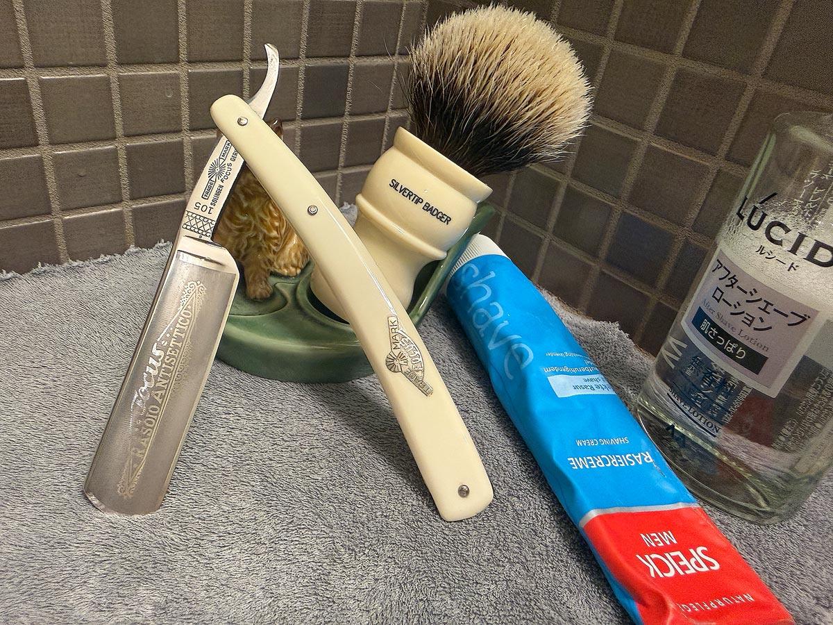 On a gray microfiber towel a straight razor with blade open about 45 degrees leans next to a bottle of aftershave and a jar of shave soap. A shave brush rests bristles-up in a green Wade ceramic pipe rest that features a brown Cairn Terrier.