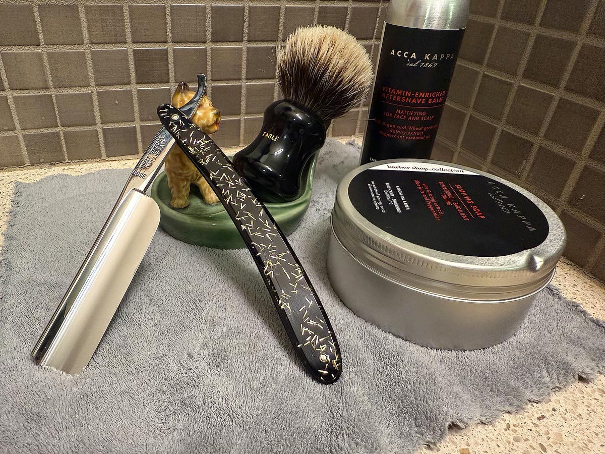 On a gray microfiber towel a straight razor with blade open about 45 degrees leans next to a bottle of aftershave and a jar of shave soap. A shave brush rests bristles-up in a green Wade ceramic pipe rest that features a brown Cairn Terrier.