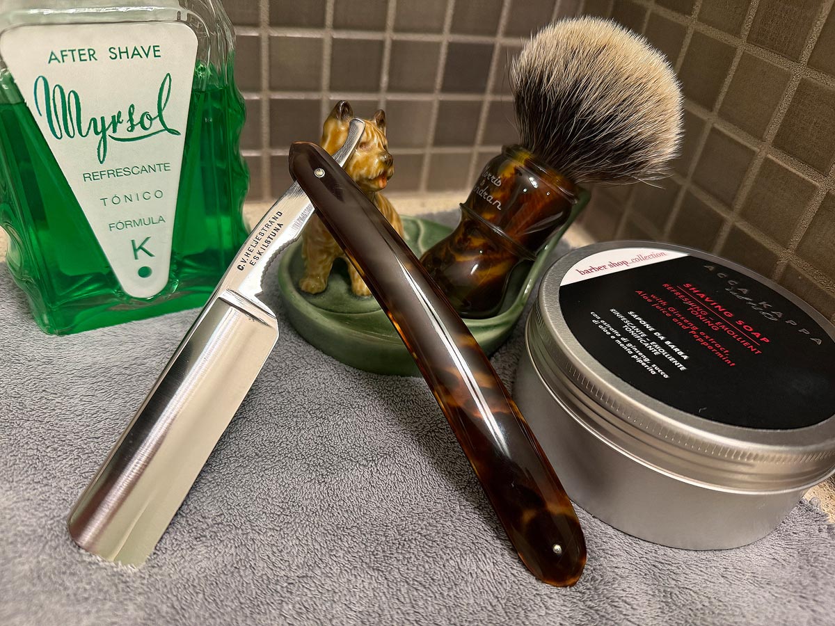 On a gray microfiber towel a straight razor with blade open about 45 degrees leans next to a bottle of aftershave and a jar of shave soap. A shave brush rests bristles-up in a green Wade ceramic pipe rest that features a brown Cairn Terrier.