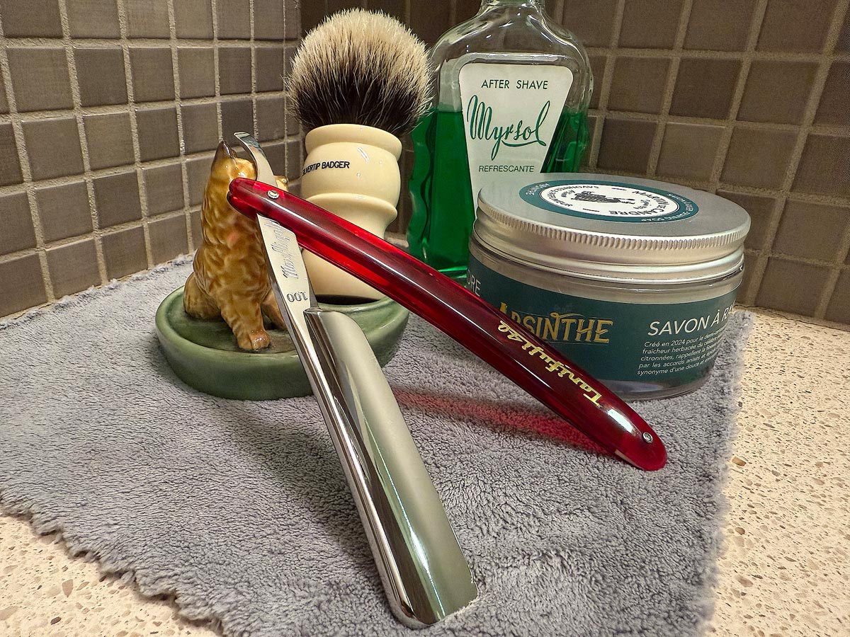 On a gray microfiber towel a straight razor with blade open about 45 degrees leans next to a bottle of aftershave and a jar of shave soap. A shave brush rests bristles-up in a green Wade ceramic pipe rest that features a brown Cairn Terrier.