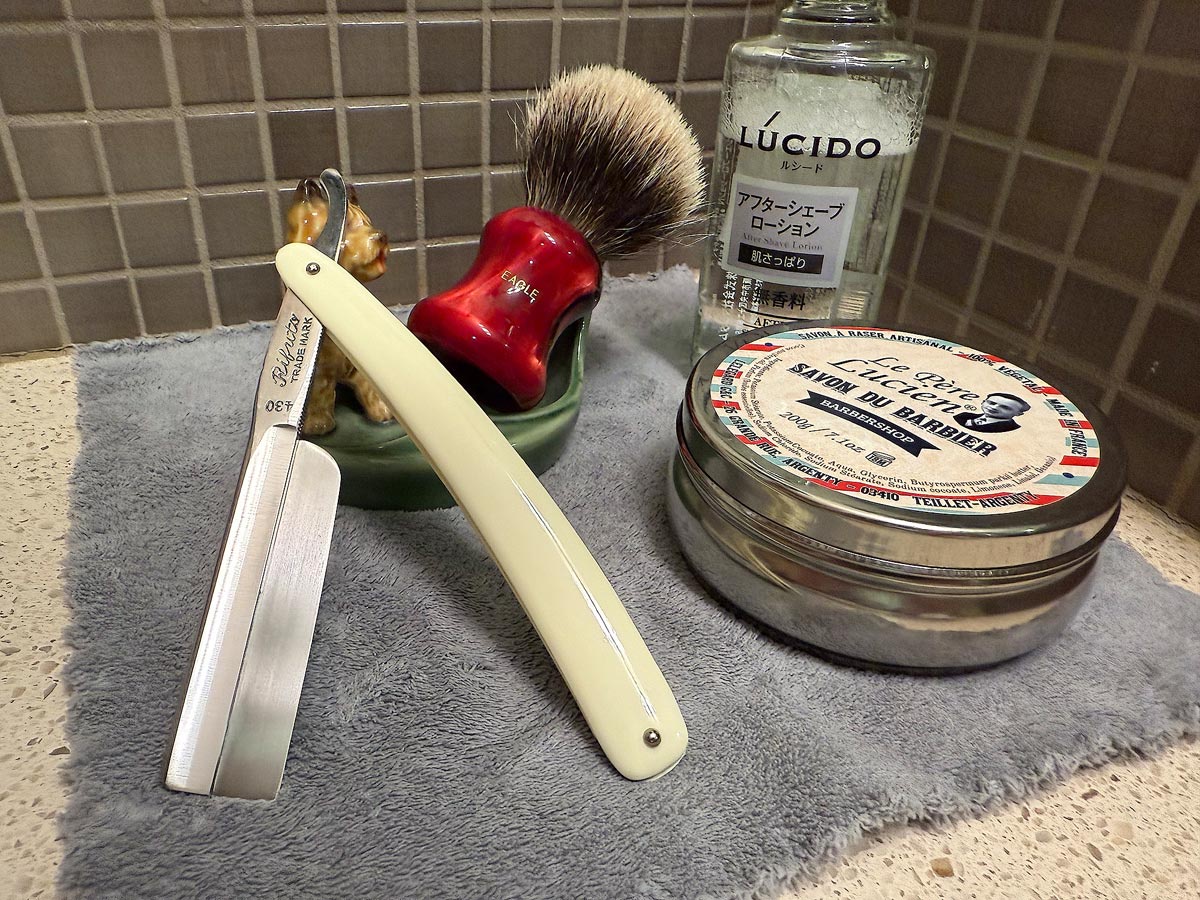 On a gray microfiber towel a straight razor with blade open about 45 degrees leans next to a bottle of aftershave and a jar of shave soap. A shave brush rests bristles-up in a green Wade ceramic pipe rest that features a brown Cairn Terrier.