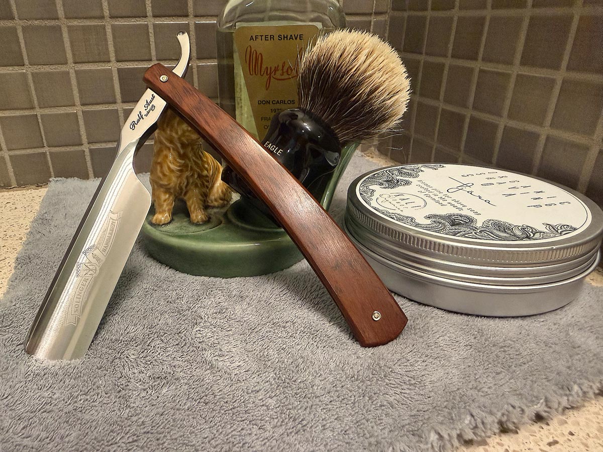 On a gray microfiber towel a straight razor with blade open about 45 degrees leans next to a bottle of aftershave and a jar of shave soap. A shave brush rests bristles-up in a green Wade ceramic pipe rest that features a brown Cairn Terrier.