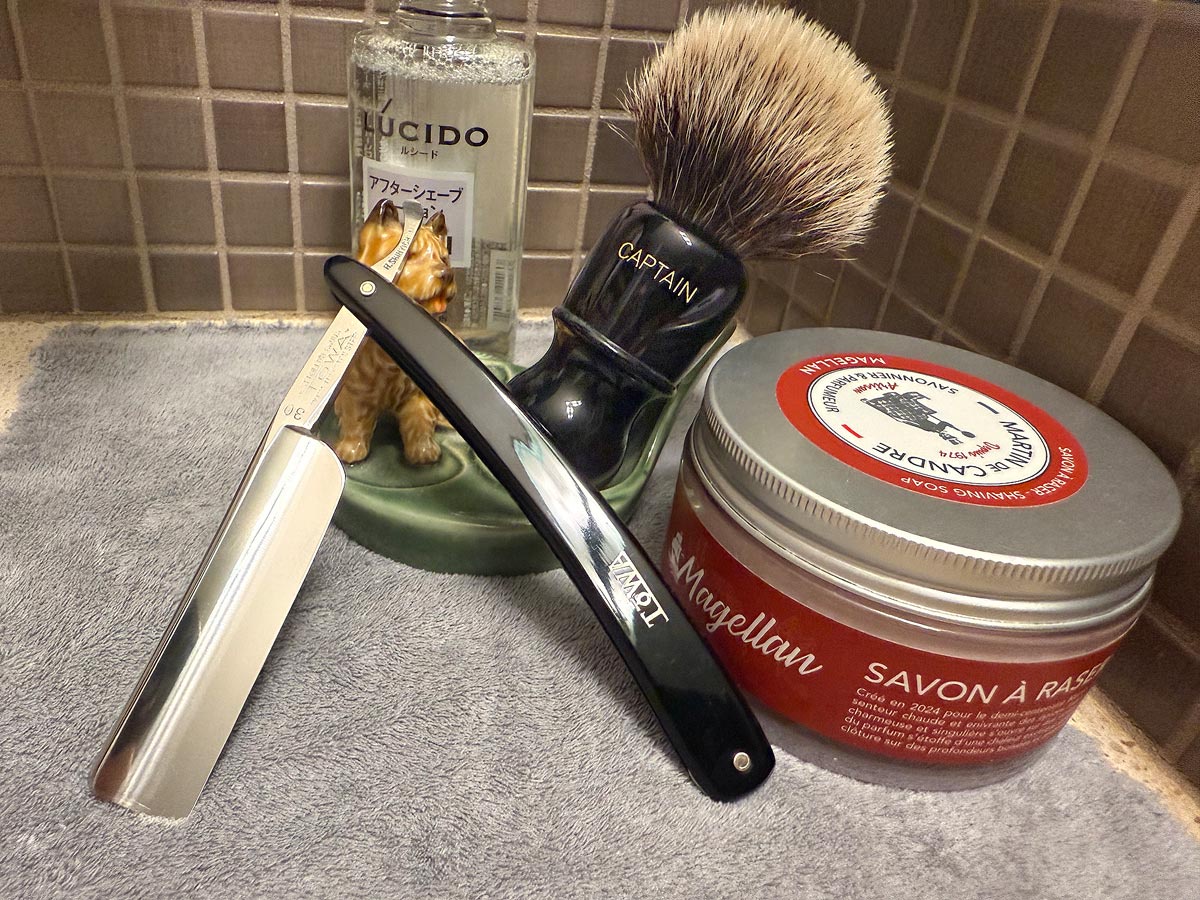 On a gray microfiber towel a straight razor with blade open about 45 degrees leans next to a bottle of aftershave and a jar of shave soap. A shave brush rests bristles-up in a green Wade ceramic pipe rest that features a brown Cairn Terrier.